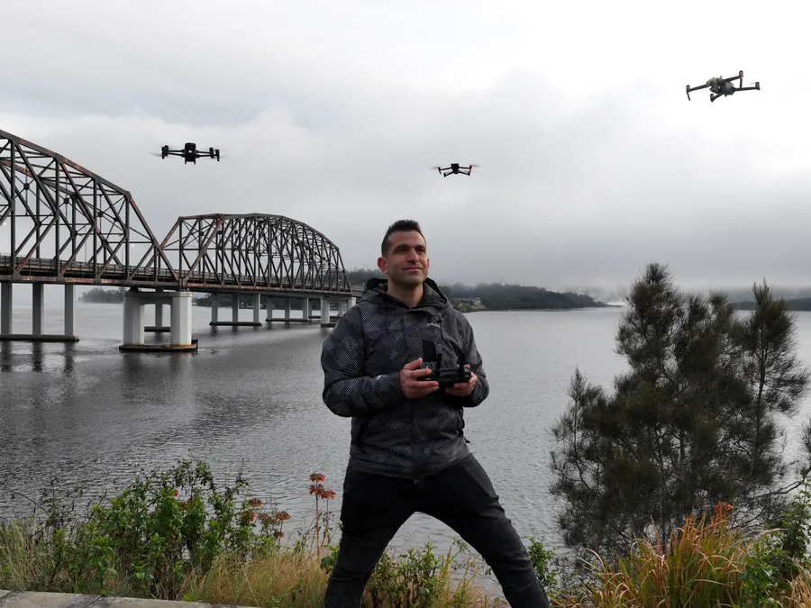 Briel Uriel on the controller with three drones in the air beside a river bridge
